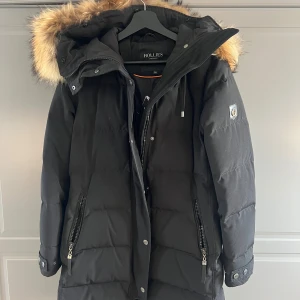 Svart dunjacka med pälskrage Hollies - Säljer en svart lång dunjacka från Hollies med stor fluffig pälskant på huvan. Jackan har dragkedjor på fickorna, knappar vid ärmslut och snygga detaljer. Perfekt för kalla vinterdagar och har en cool Arctic Expedition-tryck på insidan. Jackan är äkta med äkta päls❤️