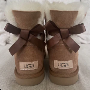 UGG boots med rosett bailey bow  - Säljer ett par klassiska UGG boots i beige mocka med fluffig vit fårull på insidan. Skorna har en söt brun rosett baktill och en platt sula. Perfekta för kalla dagar och riktigt mysiga att ha på sig. Box medföljer❣️ De har vart väl impregnerade så är i fint skick trots att de är använda, dock finns typ två fläckar som inte syns❣️Storlek 40 men passar mig med 38/39 så 38-40 borde funka❣️Pris kan diskuteras ❣️