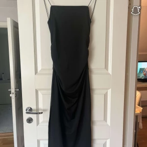 Svart långklänning med rosett och cut-out - Snygg svart långklänning med smala axelband och en cool rosettdetalj baktill som skapar en fin look. Klänningen har en elegant, rak siluett och är perfekt för dig som gillar stilrena och trendiga plagg. Materialet känns mjukt och följsamt mot kroppen.