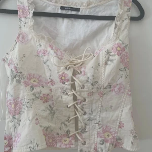 Blommig korsettopp från Gina Tricot - Supersöt korsettopp från Gina Tricot i vitt med rosa blommor och snörning framtill. Toppen har spetsdetaljer vid urringningen och är ärmlös. Perfekt för dig som vill ha en romantisk och trendig look.