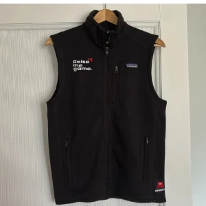 Svart fleeceväst från Patagonia - Svart fleeceväst från Patagonia med hög krage och dragkedja framtill. Västen har broderad text 'Raise the game.' på bröstet och två sidofickor med dragkedja. Perfekt för lager-på-lager och outdoor. Snygg, enkel design med diskret logga.