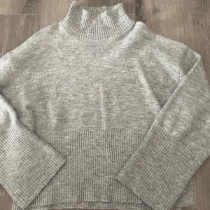 Grå stickad polotröja med ribbade detaljer - Mysig grå stickad polotröja med bred ribbad kant nertill och på ärmsluten. Hög polokrage som ger extra värme och stil. 