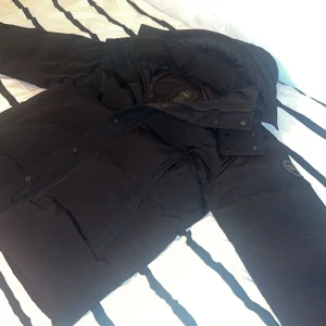 Canada Goose Jacka - Säljer en svart Canada Goose jacka i storlek L. Jackan har en klassisk pufferstil med hög krage, dragkedja och tryckknappar framtill. Ikonisk Canada Goose-logga på ärmen (Black Label). Perfekt för kalla vinterdagar och riktigt varm. För dig som behöver en fet jacka till vintern hör av dig! Öppen via byten!