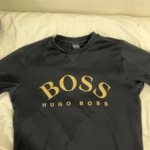 Svart sweatshirt från Hugo Boss - Svart sweatshirt från Hugo Boss med stort gult BOSS-tryck på bröstet. Klassisk rund halsringning och långa ärmar. Tröjan har en enkel och stilren design, perfekt för dig som gillar streetwear och vill ha något snyggt och bekvämt.
