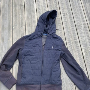 Mörkblå Cardigan/Hybridjacka från Polo Ralph Lauren - Säljer en mörkblå cardigan/hybridjacka från Polo Ralph Lauren med dragkedja och huva. Den är rätt så solskadad. Tröjan har quiltade partier framtill och på axlarna, samt ribbade muddar. Klassisk logga på bröstet och praktiska fickor. Perfekt för lager-på-lager under höst och vinter. Nypris 3000kr