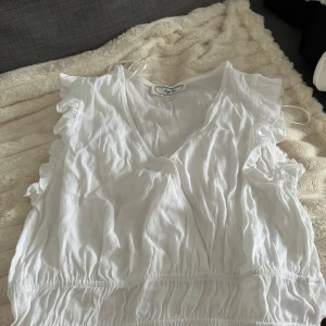 Vit blus med volang från Hippie Rose - Supersöt vit blus från Hippie Rose med v-ringning, volangdetaljer vid axlarna och resår i midjan. Blusen är ärmlös och har en luftig känsla, perfekt för varma dagar. Materialet är mjukt och lätt, vilket gör den extra skön att bära.