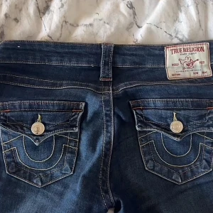 True Religion bootcut jeans blå - Säljer ett par ikoniska True Religion jeans i mörkblå denim med bootcut passform. Byxorna har låg midja, snygga kontrastsömmar och stora bakfickor med lock och silverfärgade knappar. Klassisk logga på bakfickan och patch bak i midjan. Storlek 25 💖