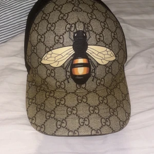 Gucci keps med bi och GG-mönster - Snygg keps från Gucci med klassiskt GG-monogram i beige och brunt. Framsidan pryds av en stor bi i gult, svart och orange. Baksidan är i svart mesh för extra ventilation. Perfekt statement-piece för dig som gillar lyxiga accessoarer. Inte så enkel få tag på och väldigt fin