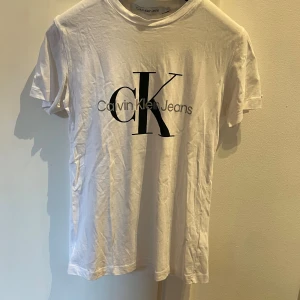 Vit t-shirt från Calvin Klein Jeans - Vit t-shirt från Calvin Klein Jeans med stor svart CK-logga och text på bröstet. Klassisk rund hals och korta ärmar. T-shirten är i mjuk bomull och har en clean, stilren look som funkar till jeans eller shorts. Perfekt för dig som gillar enkel och snygg design.
