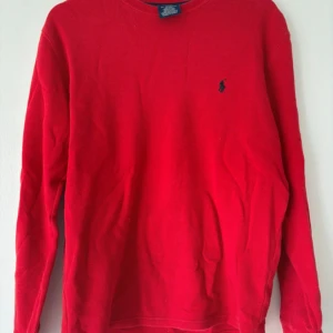 Röd långärmad tröja från Polo Ralph Lauren - Riktigt snygg röd långärmad tröja från Polo Ralph Lauren med klassisk blå broderad logga på bröstet. Tröjan är i bomull och har en enkel, stilren design med rund hals. Perfekt för dig som gillar en clean och tidlös look. Använt under jul månad 2 vintrar, använd enbart hemma. 