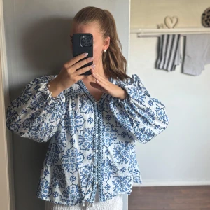 Blommig vit och blå blus med puffärm - Säljer en superfin blus med blommigt blått mönster på vit botten. Den har puffiga långärmar med resår vid handlederna, v-ringad hals och knappar framtill. Använd ett fåtal gånger och är vi fint skick🌸 superfin till semester eller middagar
