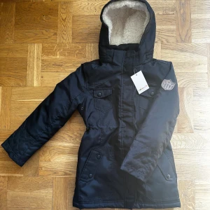 Svart vinterjacka från Colours Kids - Svart vinterjacka från Colours Kids med mysigt teddyfoder i huvan och quiltat innerfoder. Jackan har flera fickor med knappar, dragkedja framtill och en badge på ärmen. Perfekt för kalla dagar och riktigt snygg stil med praktiska detaljer.