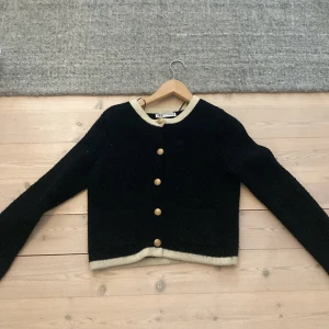 Svart kofta med guldknappar från Zara - Snygg svart kofta från Zara med kontrasterande beige kant vid hals, ärmslut och nederkant. Guldiga knappar framtill och två fickor. Långärmad och stickad i mjukt material, perfekt för lager-på-lager-stil.