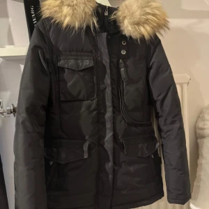 Hollies jacka - Snygg svart dunjacka från Hollies med fluffig päls runt huvan. Jackan har flera fickor, dragkedja och knappar framtill samt en märke på ärmen. Perfekt för kalla vinterdagar och riktigt stilren look.