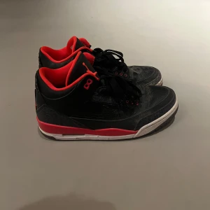 Nike Air Jordan 3 svart/röd strl 41 - Säljer ett par Nike Air Jordan 3 i svart skinn med röda detaljer och vit sula. Klassisk Jumpman-logga bak och röd insida. Snygg och ikonisk siluett med snörning och rund tå. Perfekta för dig som gillar streetwear och sneakers med attityd.