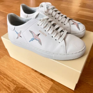 Vita sneakers från Axel Arigato med broderi - Snygga vita sneakers från Axel Arigato i skinn med broderade fåglar i rosa, blått och grått på sidorna och tån. Klassisk låg modell med snörning och rund tå. Perfekta för dig som gillar stilrena men unika detaljer. Har endast använts 2-3 gånger då de visade sig vara för små för mig, därför säljer jag dem. Originalkartong medföljer i köpet. Prissänkt från 2000-1800kr. 