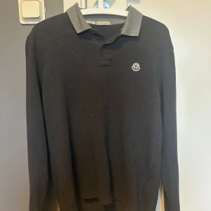 Mörkgrå Moncler pikétröja slim fit - Snygg mörkgrå långärmad pikétröja från Moncler med kontrasterande ljusgrå krage och ärmslut. Slim fit-modell med klassisk pikéknäppning. STRL S skriv vid minsta fundering!