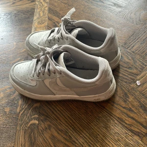 Nike Air Force 1 i ljusgrått skinn - Nike Air Force 1 sneakers i ljusgrått skinn med detaljer i mocka på hälen. Klassisk låg modell med snörning och perforerad tå för extra ventilation. Snygg och tidlös design som passar till det mesta. Swoosh-logga på sidan och broderad Nike Air på baksidan.