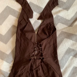 Brun långklänning med snörning från Bershka - Säljer en brun långklänning från Bershka i storlek L. Klänningen har snygg snörning framtill, dragkedja och breda axelband. Materialet är mjukt och känns som bomull, perfekt för dig som gillar en cool och enkel stil. Passformen är normal och klänningen är ärmlös.