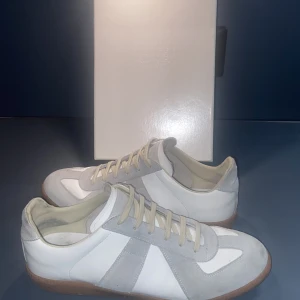 Maison Margiela Gats  - Maison Margiela Gats sneakers i vitt skinn och ljusgrå mocka med klassisk gummisula. Snygg retrodesign med rund tå och snörning. Perfekta för dig som gillar minimalistisk och stilren look. Kommer med originalkartong.