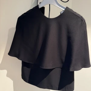 Svart omlott blus med cape-detalj - Stilren svart blus med cape-liknande lager över axlarna och rund halsringning. Toppens flowiga tyg ger en cool och elegant siluett, perfekt för att sticka ut. Passar dig som gillar minimalistisk men unik stil.