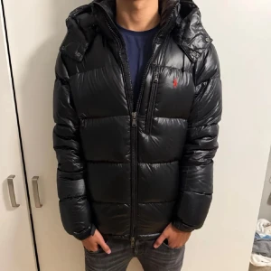 Svart Ralph Lauren glansig Vinterjacka - Polo Ralph Lauren glossy svart jacka med en röd häst | Storlek L passar runt 180 cm | Inga stora skador | 2 pyttensmå hål ingen märker finns 🥶| Kan skicka bild på den andra om du vill | Äkta nypris runt 6000kr | Hör av dig 🤙 