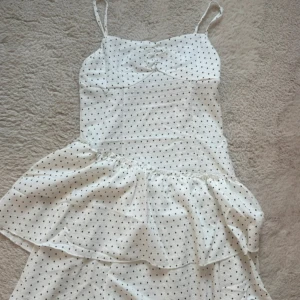 Vit prickig kortklänning med volang - Supersöt vit kortklänning med svarta prickar och volang nertill. Klänningen har smala axelband och en feminin passform, perfekt för varma dagar. Lätt och luftig, med en söt detalj framtill vid bysten.