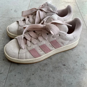 Adidas Campus sneakers i ljusrosa mocka - Snygga Adidas Campus sneakers i ljusrosa mocka med klassiska tre ränder i rosa på sidorna. Skorna har breda matchande skosnören, rund tå och platt sula. Adidas logga på plös och häl. Perfekta för dig som gillar en clean och trendig look.