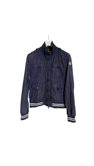 Marinblå vindjacka från Moncler - Snygg marinblå vindjacka från Moncler med ribbad krage och muddar med vita ränder. Jackan har fyra fickor framtill med knappar, dragkedja och Moncler-logga på ärmen. 