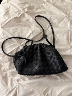 Svart flätad axelväska i skinn - Snygg svart axelväska med flätad design och justerbar rem. Väskan är tillverkad i mjukt skinn och har en praktisk öppning med dragsko. Perfekt storlek för det viktigaste och ger en trendig touch till din outfit.