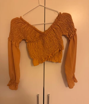 Gul croppad blus med smock - Trendig gul blus med smockad överdel och långa ärmar. Offshoulder-modell med volangdetaljer och croppad passform. Perfekt för dig som vill sticka ut med en färgglad och unik topp.