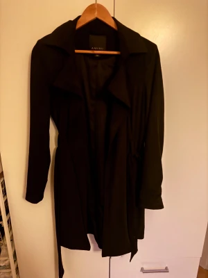 Svart trenchcoat från Amisu - Snygg svart trenchcoat från Amisu med klassisk krage och bälte i midjan. Jackan har långa ärmar och en stilren, tidlös design som passar perfekt till lager-på-lager-look. Materialet känns lätt och smidigt, vilket gör den enkel att bära under vår och höst.