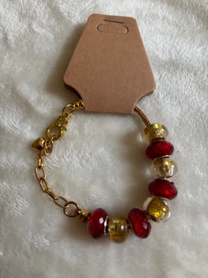 Guldarmband med röda och glittriga pärlor - Snyggt armband i guldfärgad metall med stora röda och guldglittriga pärlor. Armbandet har en kedja med hjärtberlock och karbinlås. Perfekt accessoar för att lyfta din outfit med lite extra färg och bling.