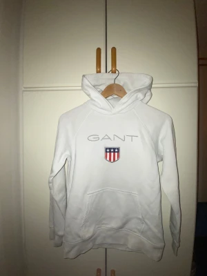 Vit hoodie från GANT med logga - Vit hoodie från GANT med broderad logga och emblem på bröstet. Tröjan har huva, långärmad design och en stor magficka framtill. Materialet är mjuk bomull och passformen är normal. Perfekt för dig som gillar klassisk och clean streetstyle. Säljer den pågrund av att den är liten på mig