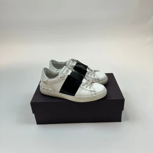 Valentino Open sneakers vit/svart - Snygga Valentino Open sneakers i vitt skinn med bred svart rem över mitten. Klassisk rund tå och platt sula, samt ikoniska nitar på hälen. Perfekt för dig som gillar stilrena och lyxiga sneakers med en edgy touch. Storlek 41