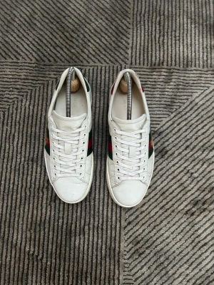 Gucci Ace vita skor - Gucci Ace sneakers i vitt skinn med klassiska gröna och röda ränder på sidorna och en broderad guldfärgad bi. Skorna har snörning, platt sula och rund tå. Perfekta för dig som vill ha en lyxig och stilren sneaker med ikoniska detaljer.