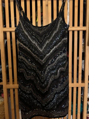 Svart glittrig paljettklänning - Supersnygg svart cocktail klänning med tunna axelband och glittriga paljetter i ett sicksackmönster. Klänningen har en figurnära passform och är perfekt för dig som vill sticka ut med en festlig look.