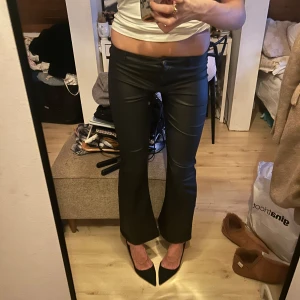 Svarta bootcut byxor i skinnimitation - Säljer ett par svarta bootcut mid waisted byxor i skin nimitation med låg midja. Snyggt glansigt material och klassisk passform som sitter tajt upptill och släpper vid benen. Perfekt för dig som gillar en edgy och trendig look. Perfekt för fest och till vardags. En favorit & strechiga