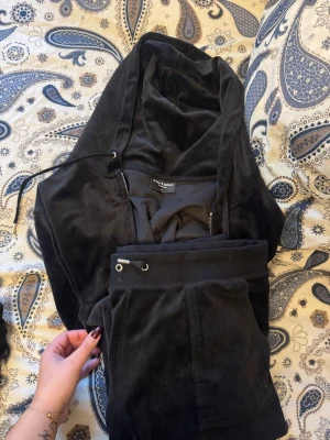 Svarta mjukisbyxor från Juicy Couture - Svarta mjukisbyxor samt koftan från Juicy Couture i mjuk sammet med snörning i midjan och fickor med silverfärgade knappar. Klassisk logga på fickan och avslappnad passform, perfekta för chill dagar. Snyggt och bekvämt val för dig som gillar ikonisk streetstyle.