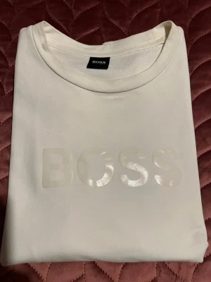 Vit sweatshirt från Hugo Boss - Snygg vit sweatshirt från Hugo Boss med BOSS-logga i relief på bröstet och längs ärmarna. Tröjan har rund hals, ribbade muddar och är tillverkad i mjuk bomull. Perfekt för dig som gillar stilrena och exklusiva plagg med diskreta detaljer.