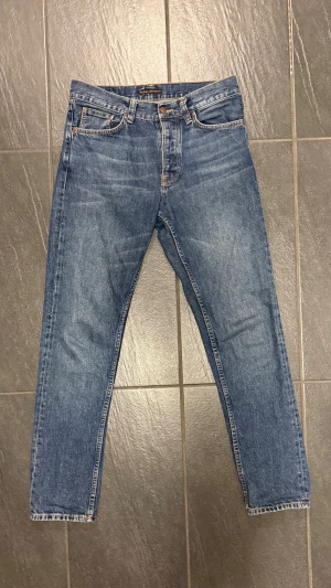 Blå jeans från Nudie Jeans, storlek 30/32 - Snygga blå jeans från Nudie Jeans i klassisk straight fit med fem fickor och orangea sömmar. Modellen har normal passform och raka ben, tillverkade i slitstarkt denim. Perfekt för dig som gillar tidlös stil och vill ha ett par jeans som funkar till allt. 