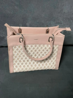 Handväska från Ulrika Sweden i rosa - Snygg handväska från Ulrika Sweden i ljusrosa syntet med flätad beige frontpanel. Väskan har silverfärgade metalldetaljer, dubbla handtag och dragkedja upptill. Insidan är fodrad med mönstrat tyg och har flera fack för smart organisering.