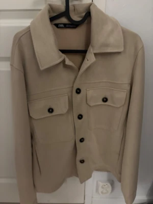 Beige mocka overshirt från Zara - Snygg beige overshirt från Zara i mjuk mocka-look med klassisk krage och svarta knappar framtill. Två stora bröstfickor med lock och knappdetalj ger en cool och stilren vibe. Perfekt att slänga över en hoodie eller t-shirt för en chill look.