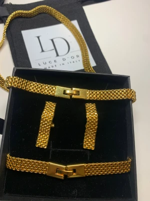 Guldfärgat smyckeset från Luce D'Oro - Snyggt smyckeset från Luce D'Oro med halsband, armband och örhängen i guldfärgad metall. Setet har en modern, fyrkantig länkdesign och stilrena spännen. Perfekt för dig som gillar statement-accessoarer med italiensk känsla.