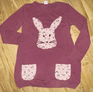 Rosa klänning med kaninmotiv Lindex - Supersöt rosa långärmad klänning från Lindex Kids med ett stort kaninansikte i blommigt tyg framtill och matchande blommiga fickor. Perfekt för dig som gillar gulliga detaljer och mjuka material.