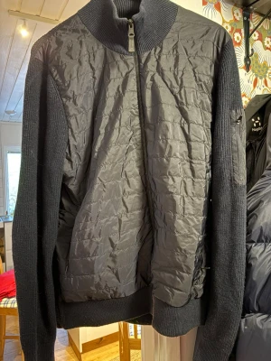 Svart quiltad bomberjacka J.Lindeberg - Snygg svart bomberjacka från J.Lindeberg med quiltad framsida och stickade ärmar. Jackan har hög krage, dragkedja framtill och en cool logga på ärmen. Materialmix av polyester och stickad ull ger både stil och komfort. Perfekt för dig som gillar streetwear med premiumkänsla.