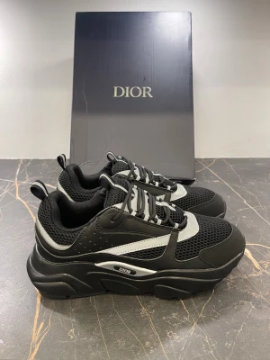 Svarta Dior sneakers med silvriga detaljer - Chunky svarta Dior sneakers med silvriga detaljer och mesh-paneler. Snyggt mönstrad sula, platt och bred för extra komfort. Skorna har snörning och reflekterande inslag som ger en cool vibe. Materialmix av skinn och syntet, med tydlig Dior-logga på sidan.