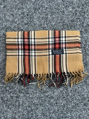 Rutig ullhalsduk från Burberry - Klassisk rutig halsduk från Burberry i 100% lammull. Beige bas med svarta, vita och röda ränder samt fransar i ändarna. Perfekt accessoar för att lyfta din outfit.
