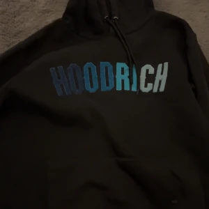 HOODRICH tröja - Svart hoodie från HOODRICH i storlek S. Stor HOODRICH-logga i blått och vitt framtill, klassisk känguruficka och justerbar huva med snören. Mjuk bomullskänsla och relaxed fit, perfekt för streetwear-stil.