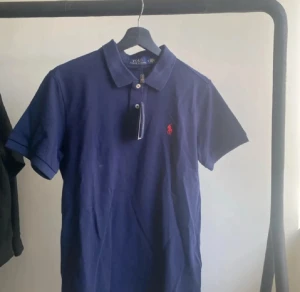 Marinblå pikétröja Polo Ralph Lauren - Snygg marinblå pikétröja från Polo Ralph Lauren i storlek S. Klassisk krage, korta ärmar och röd broderad logga på bröstet. Tröjan är gjord i mjuk bomull och har knappar vid halsen. Perfekt för en chill och stilren look.